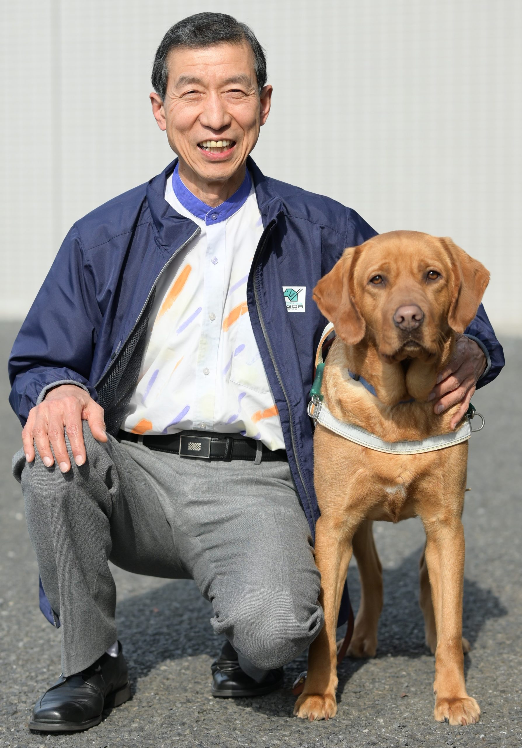 A smiling Seishi kneels next to a golden labrador in harness